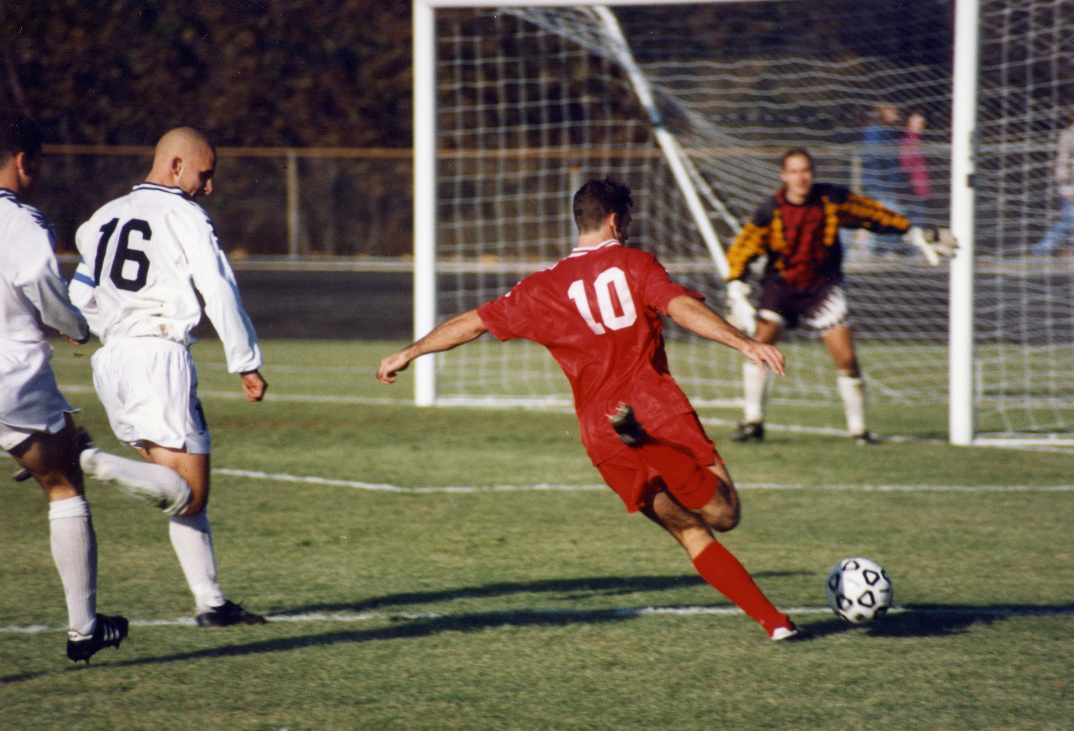 A young footballer striking the ball toward goal during a competitive club match, defender and goalkeeper in frame