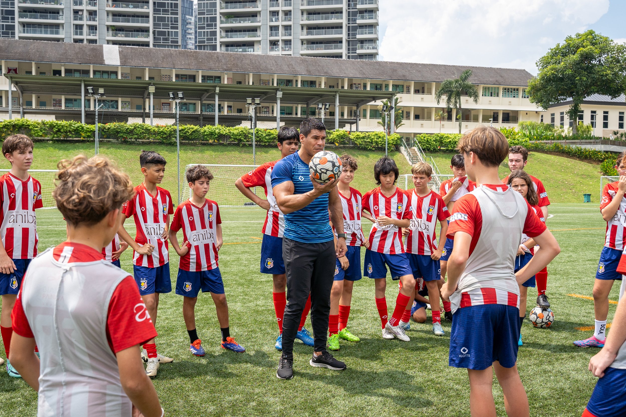 A youth football coach instructing a squad of young players during a training session on a pitch in an Asian city