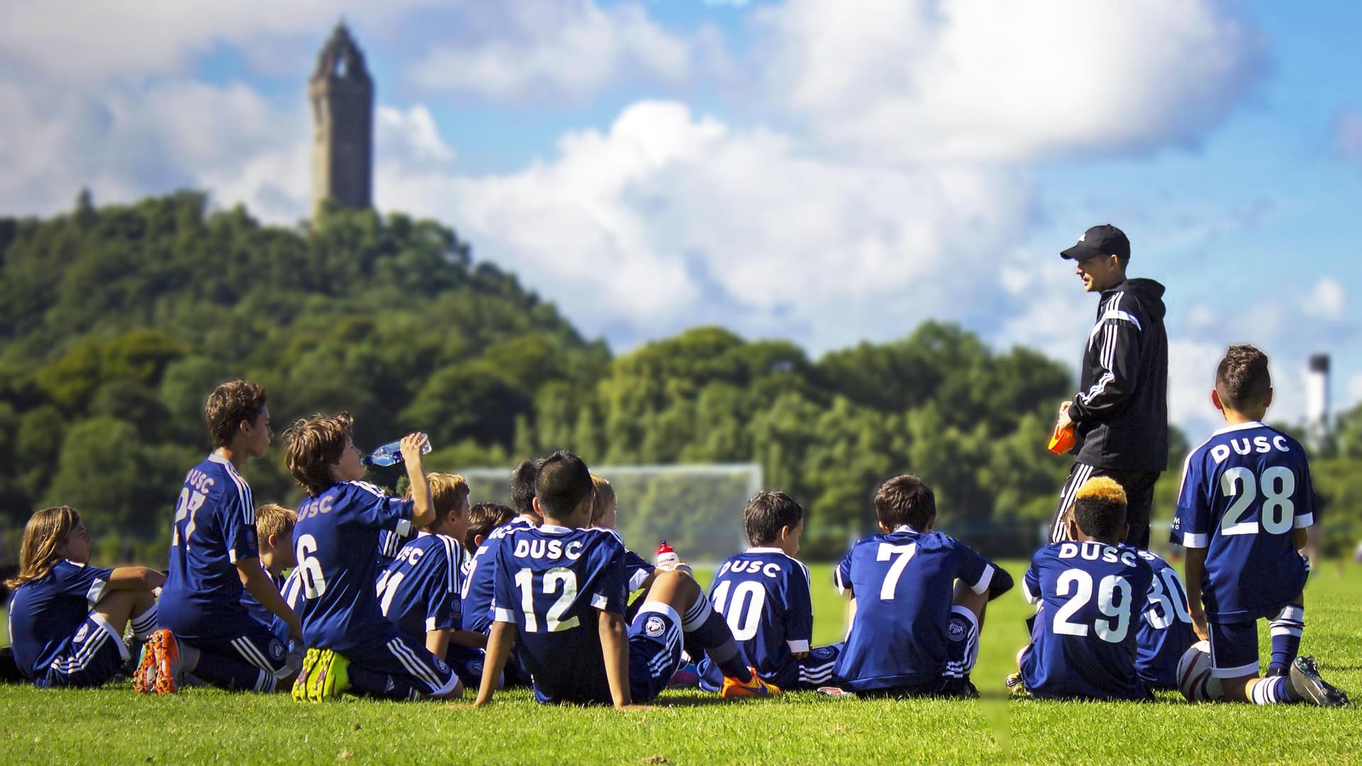 A youth football team in blue kit sitting with their coach on a grass pitch while on an international sports tour abroad