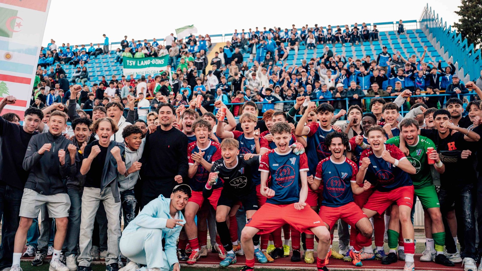 A youth football team celebrating together in a European stadium with international flags and a full crowd during a football tour