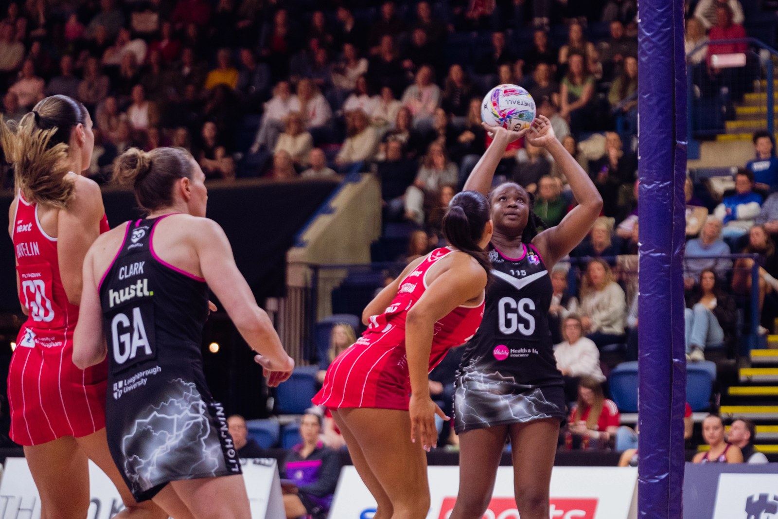 Netball players in club bibs competing at the post during a live Netball Super League match in England