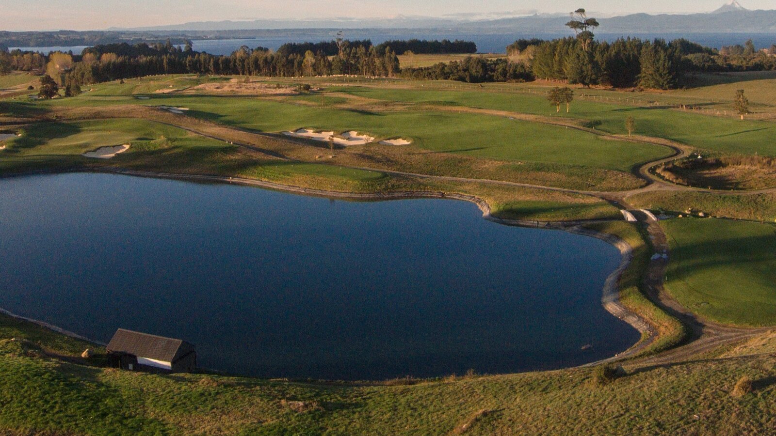Patagonian golf course at sunset with fairways wrapped around a lake, forest and distant snow-capped Andes peaks