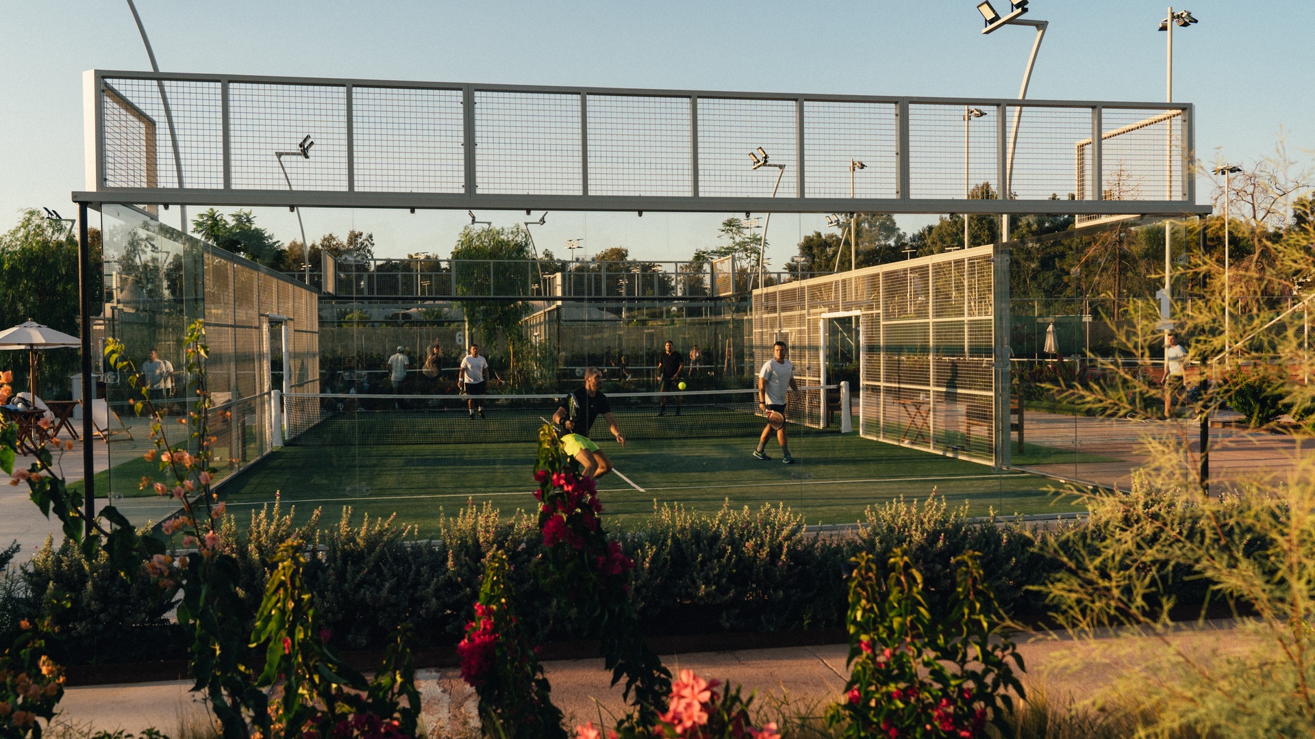 Outdoor glass-walled padel court at golden hour in a Mediterranean setting with players mid-match