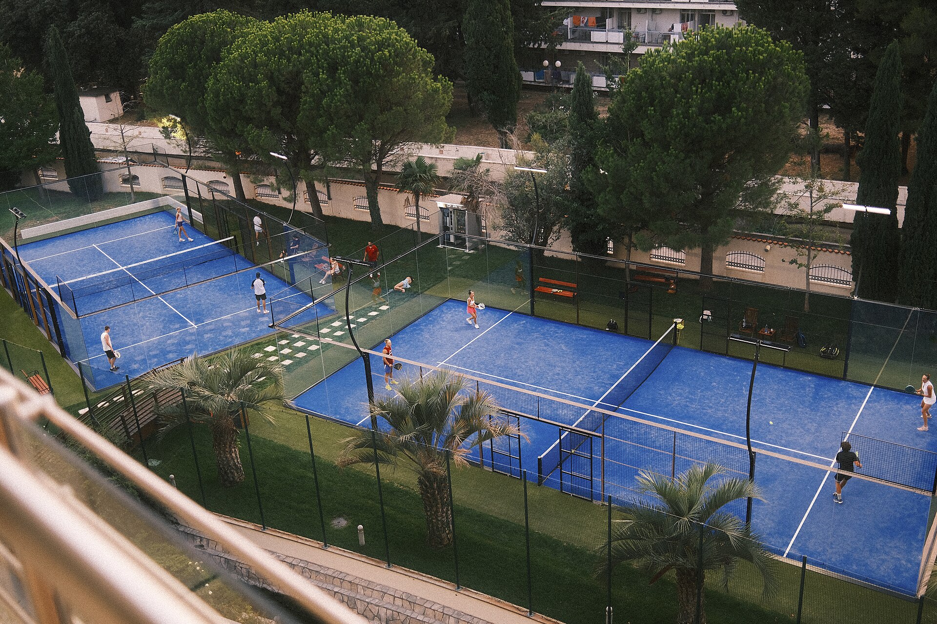 Players in a padel match on blue outdoor courts surrounded by trees