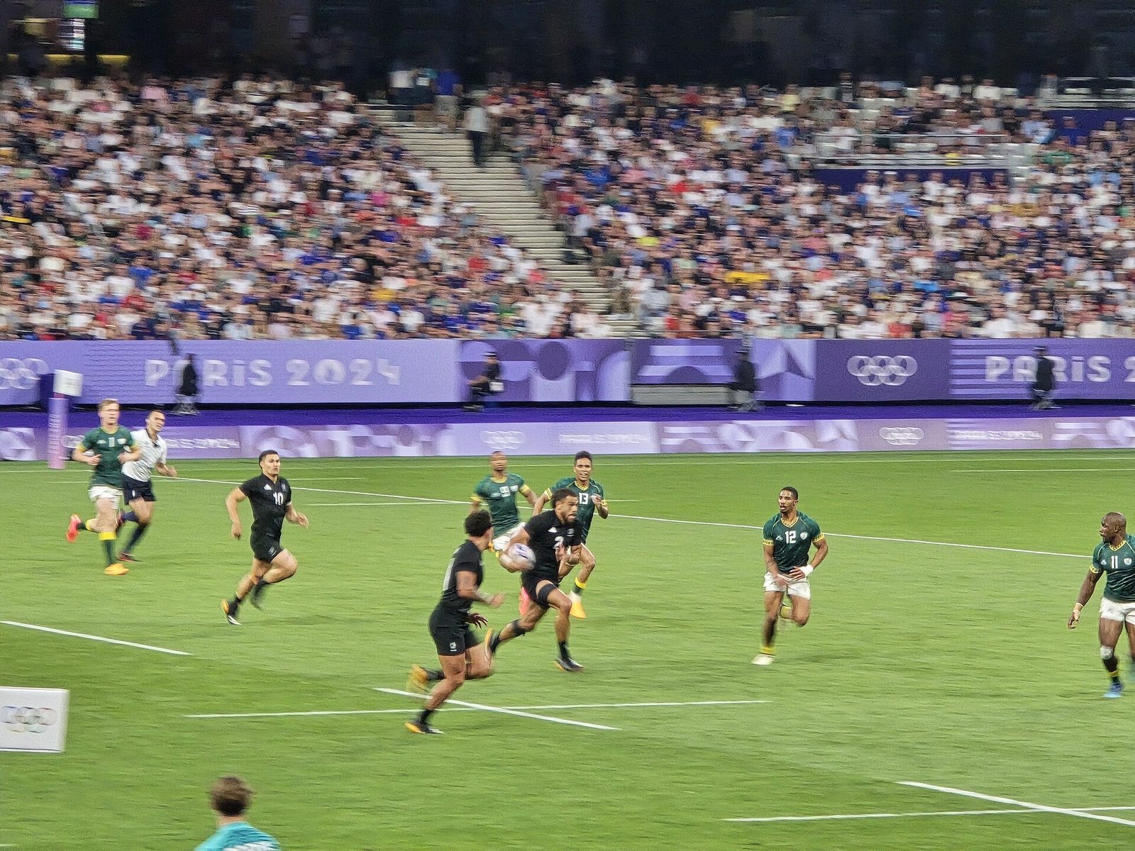 Professional rugby sevens players in black and green sprinting with the ball in a packed stadium